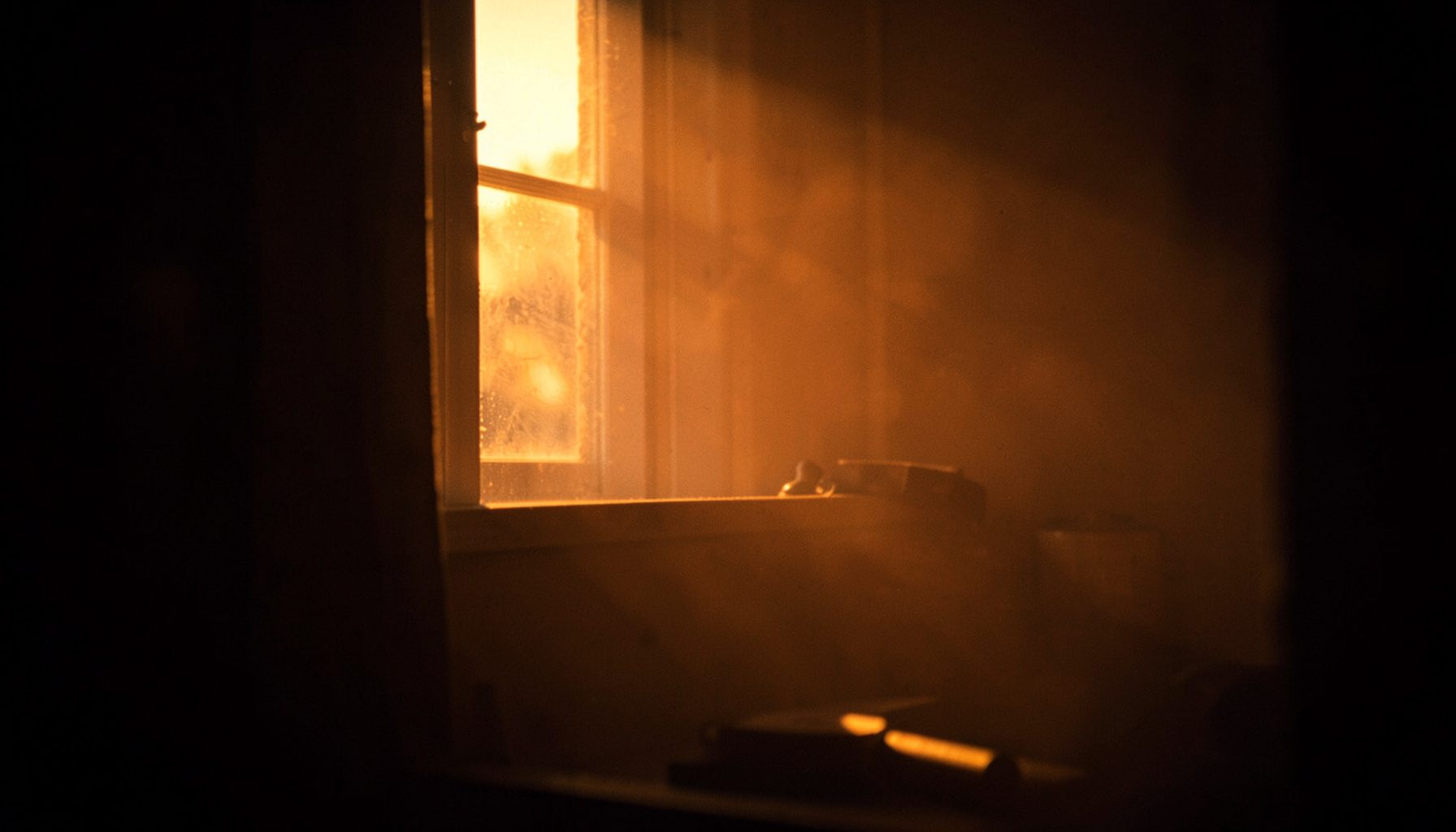 A dark cinematic still with warm golden-hour light filtering through a window onto a quiet scene